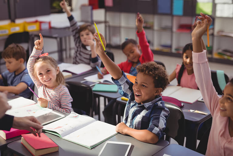 A classroom of 7 students raising their hands. In front of the students are open notebooks.