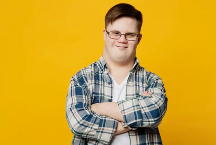 A teen with down's syndrome wearing a blue checkered shirt over a white shirt. He is wearing a smile with his arms crossed.