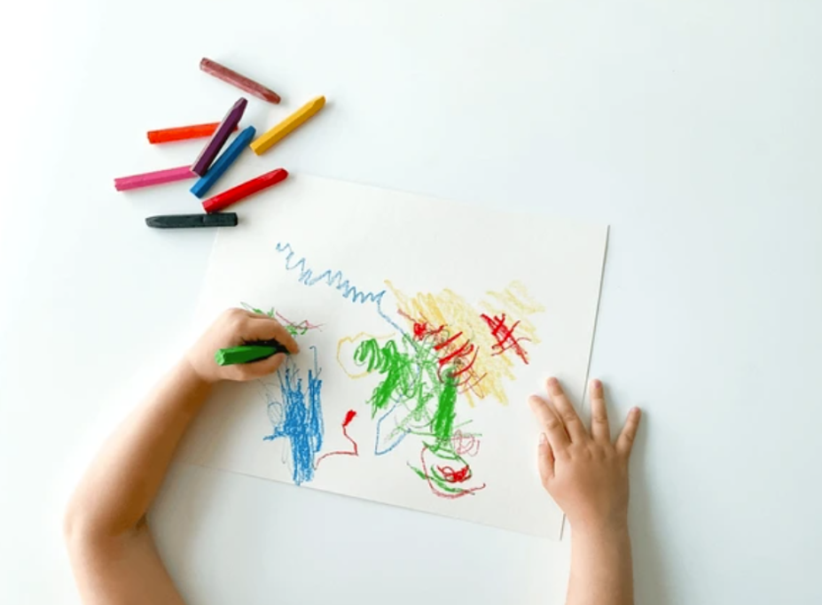 A child creates a drawing with blue, green, red, and yellow scribbles on a white paper against a white table. 8 crayons are on the side of the drawing. 