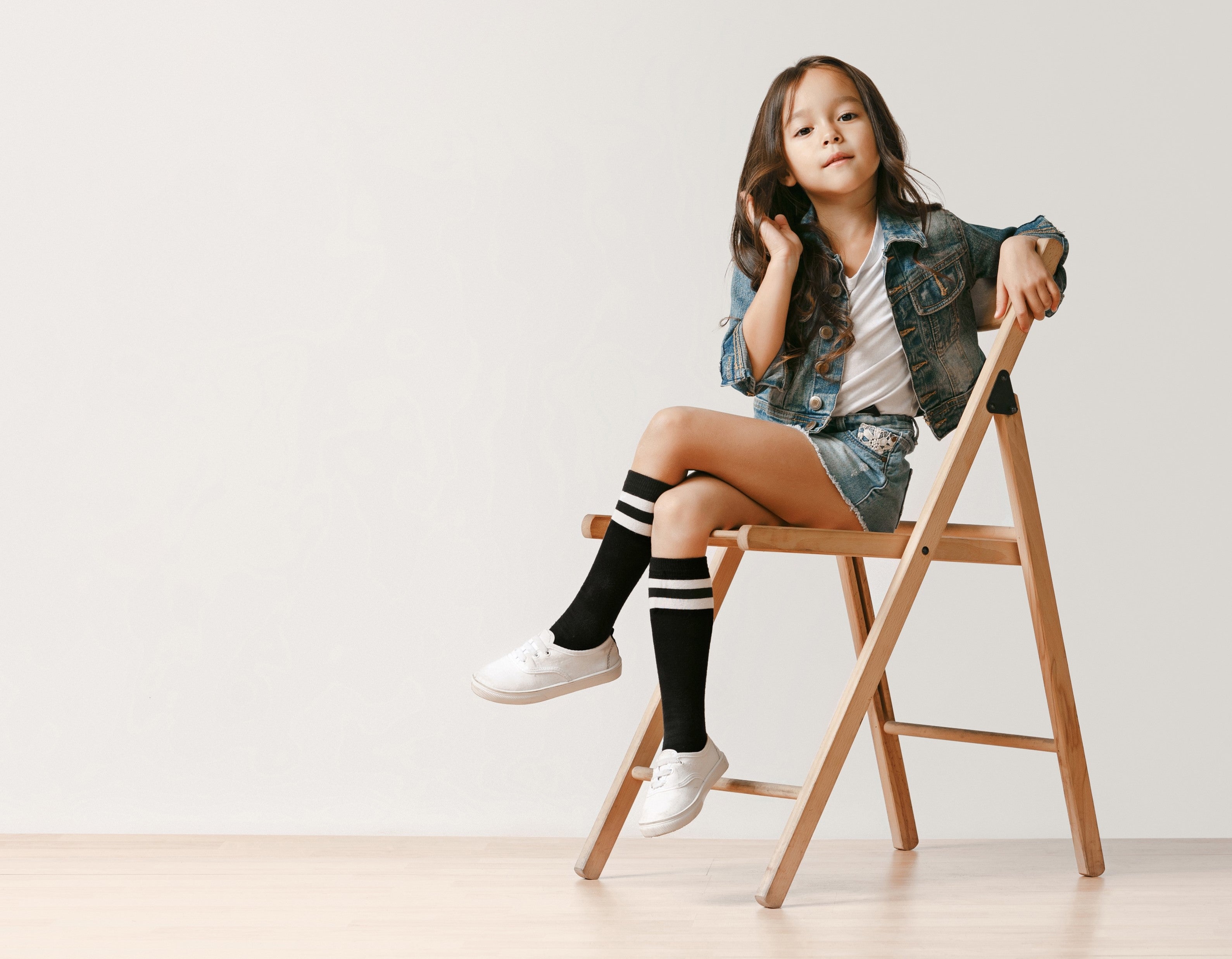 A girl sits on a wooden chair dressed in a denim jacket and skirt with a white shirt, black thigh high socks with a 2 white stripes at the top and canvas sneaker