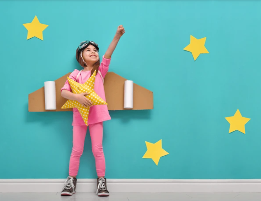 A girl in a pink top and leggings stretches her left fist towards the sky. She is dressed as a pilot wearing a 1930s fighter pilot hat, googles on top of the hat, and cardboard and styrofoam airplane wings on her back. She is holding a yellow star plush toy. The background is a blue wall with white trim featuring 4 large yellow stick-on stars.