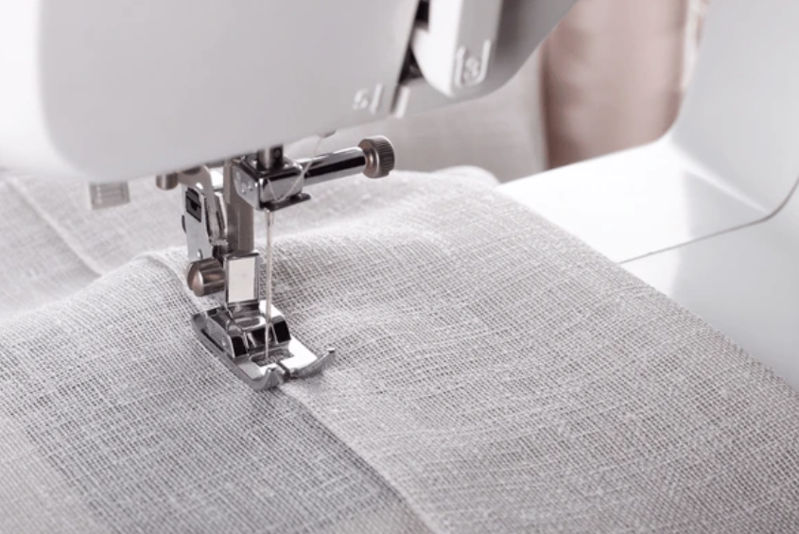 A close up of a sewing machine creating a stitch on light gray linen fabric