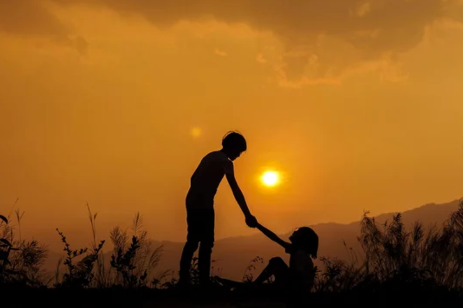 One child reaches out their hand to help another child who is sitting in the grass. The background is of a sunset