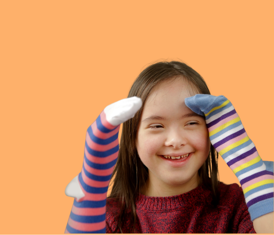 A girl with Down's syndrome in a red sweater smiles while wearing colorful striped sock on her hands against an orange background