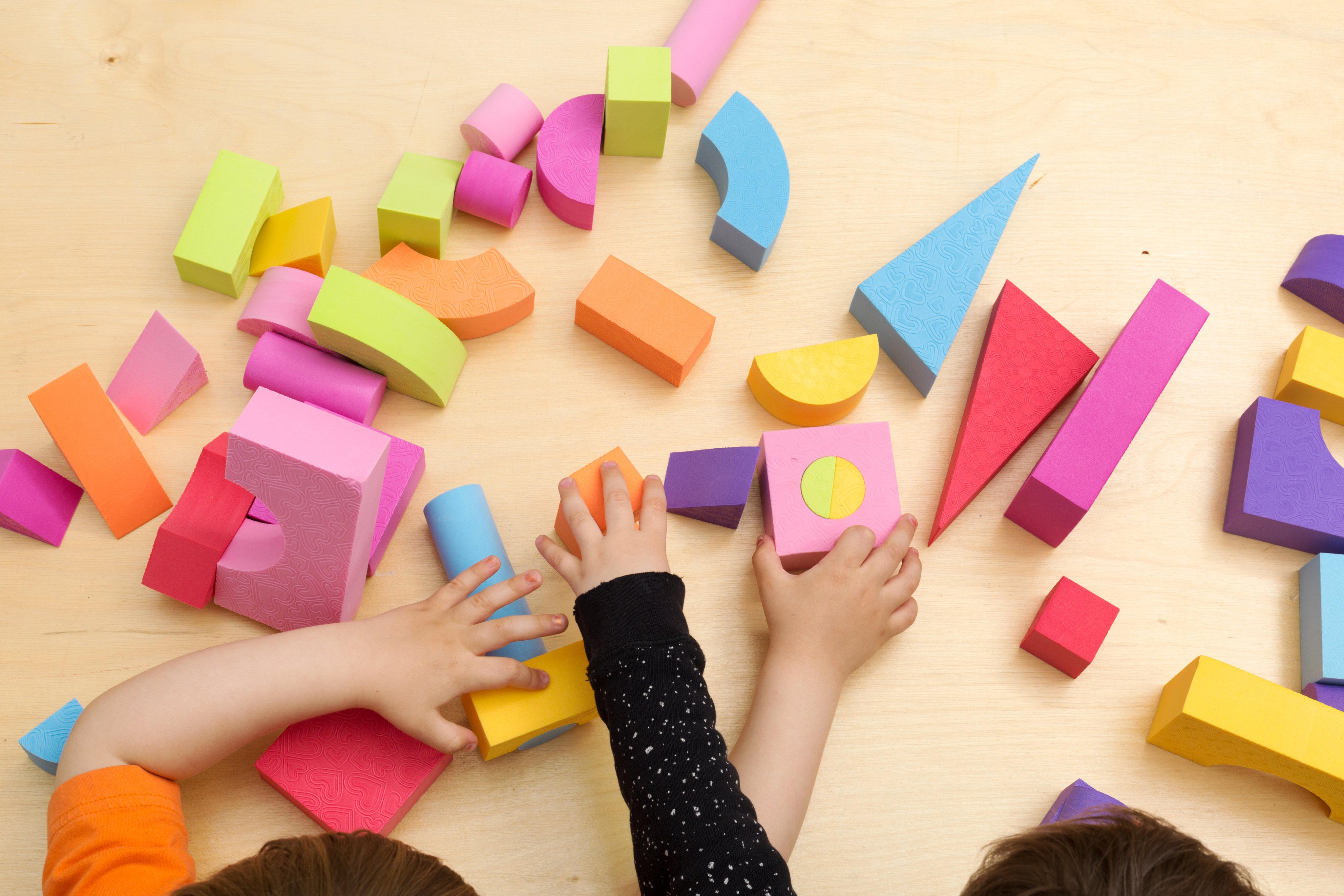 Several childrens' hands are touching and rearranging colorful building blocks. The blocks include shapes such as triangles, arches, cylinders, and cubes.