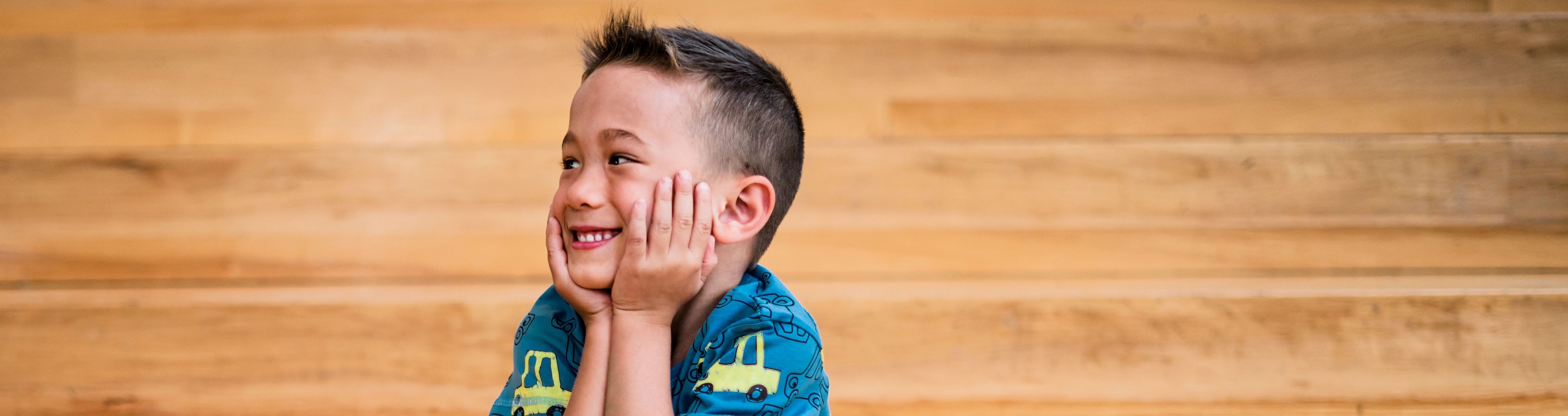 A boy looks to the left with a smile on his face and both his hands on his cheeks. He is wearing a blue shirt and sitting against a light wood panel background.