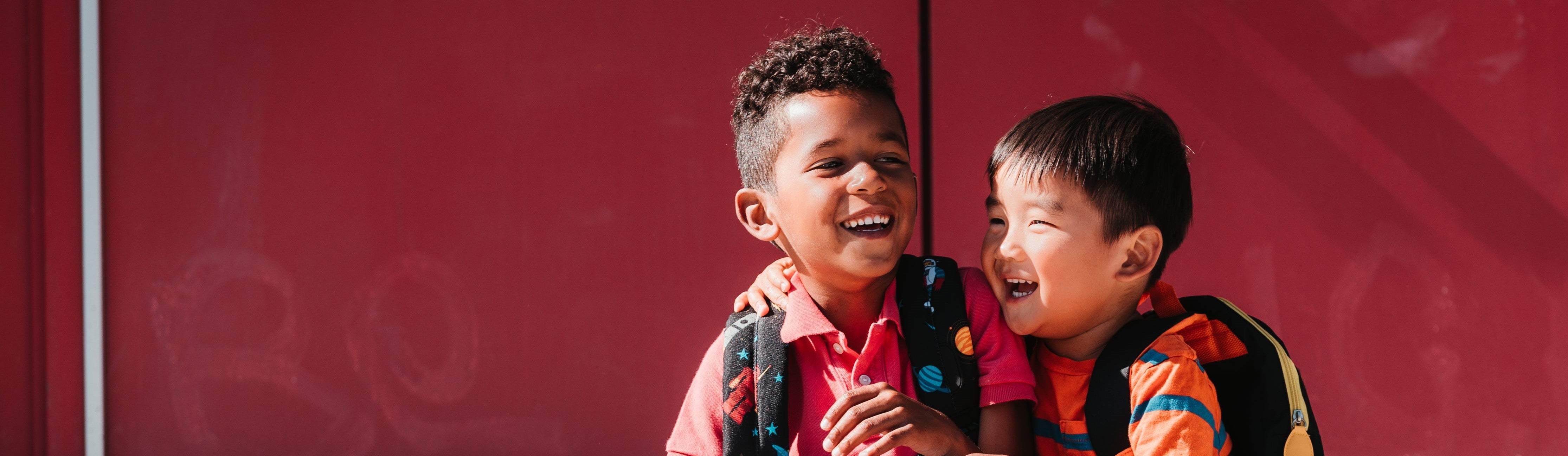 Two school boys with their arms around each other laughing together in front of a red door